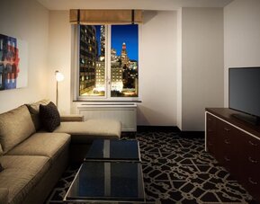 Living area with sofa, coffee table, television, and city view through window at Hilton Brooklyn New York.