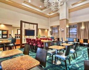 Dining area with multiple tables and chairs, patterned carpet, chandelier lighting, and breakfast counter with beverage station at Marriott SpringHill Suites Naperville/Warrenville.