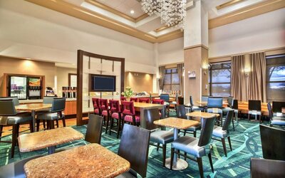 Dining area with multiple tables and chairs, patterned carpet, chandelier lighting, and breakfast counter with beverage station at Marriott SpringHill Suites Naperville/Warrenville.