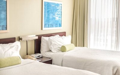 Two double beds with white linens and green bolster pillows, shared nightstand with phone, wall art, and large window with sheer curtains at Marriott SpringHill Suites Naperville/Warrenville.
