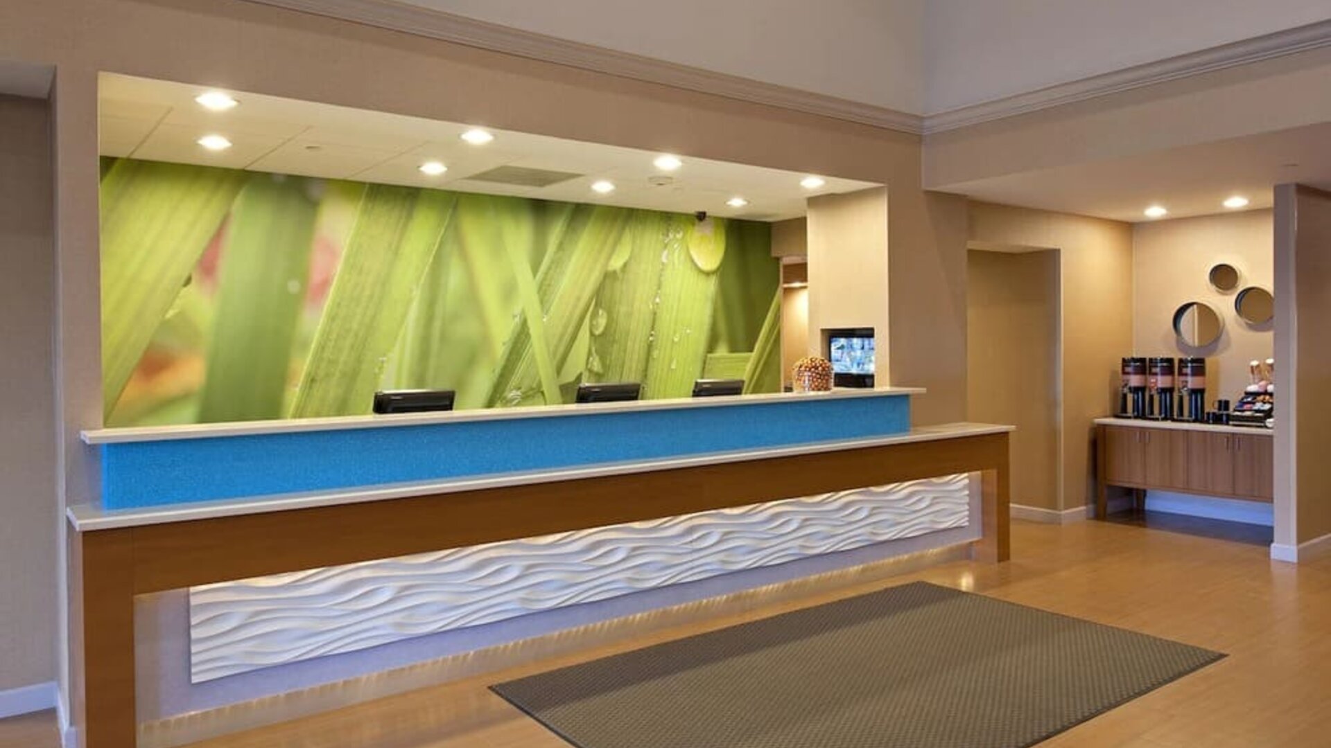 Front desk area with textured counter, green nature mural backdrop, and beverage station to the side at Marriott SpringHill Suites Naperville/Warrenville.