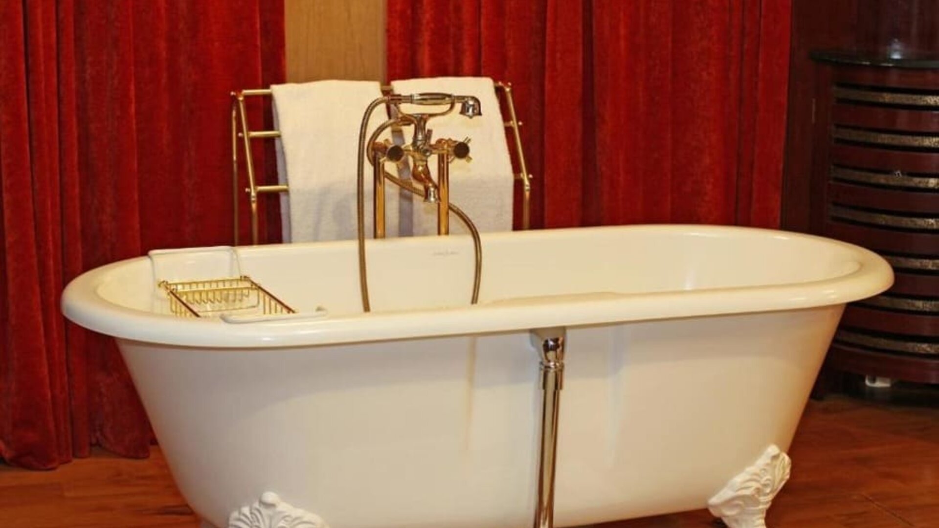 Close-up of vintage clawfoot tub with handheld shower, brass taps, and red curtain backdrop at Hôtel Espérance.