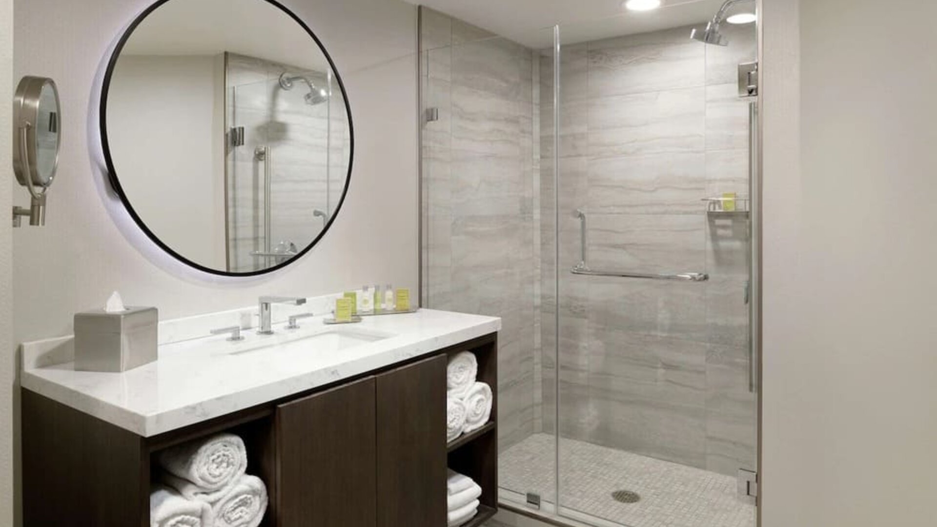 Modern bathroom with glass walk-in shower, large round mirror, vanity, and folded towels at DoubleTree By Hilton Los Angeles Downtown.