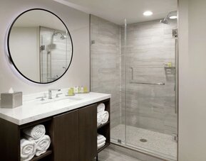 Modern bathroom with glass walk-in shower, large round mirror, vanity, and folded towels at DoubleTree By Hilton Los Angeles Downtown.