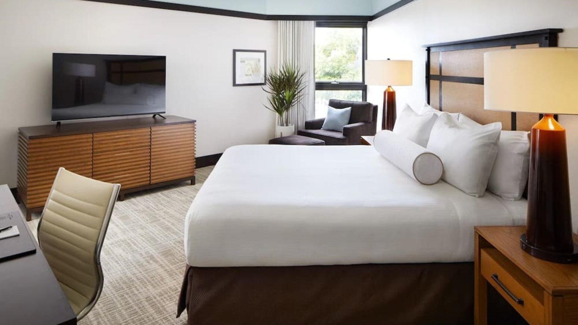 Guest room with king bed, TV console, seating chair, and large window bringing in natural light at The Ambrose Hotel Santa Monica.