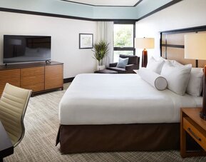 Guest room with king bed, TV console, seating chair, and large window bringing in natural light at The Ambrose Hotel Santa Monica.