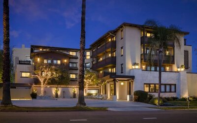 Exterior view at dusk with lit facade, palm trees, and entrance canopy along the street at The Ambrose Hotel Santa Monica.