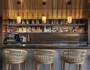 Sleek bar with backlit shelves, bottle display, pendant lighting, and woven bar stools at AC Hotel By Marriott Beverly Hills.