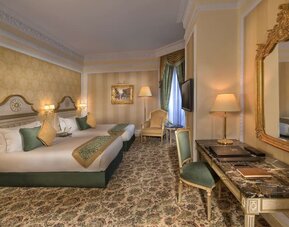 Twin bedroom with two beds, patterned carpet, armchair seating, and wall-mounted television