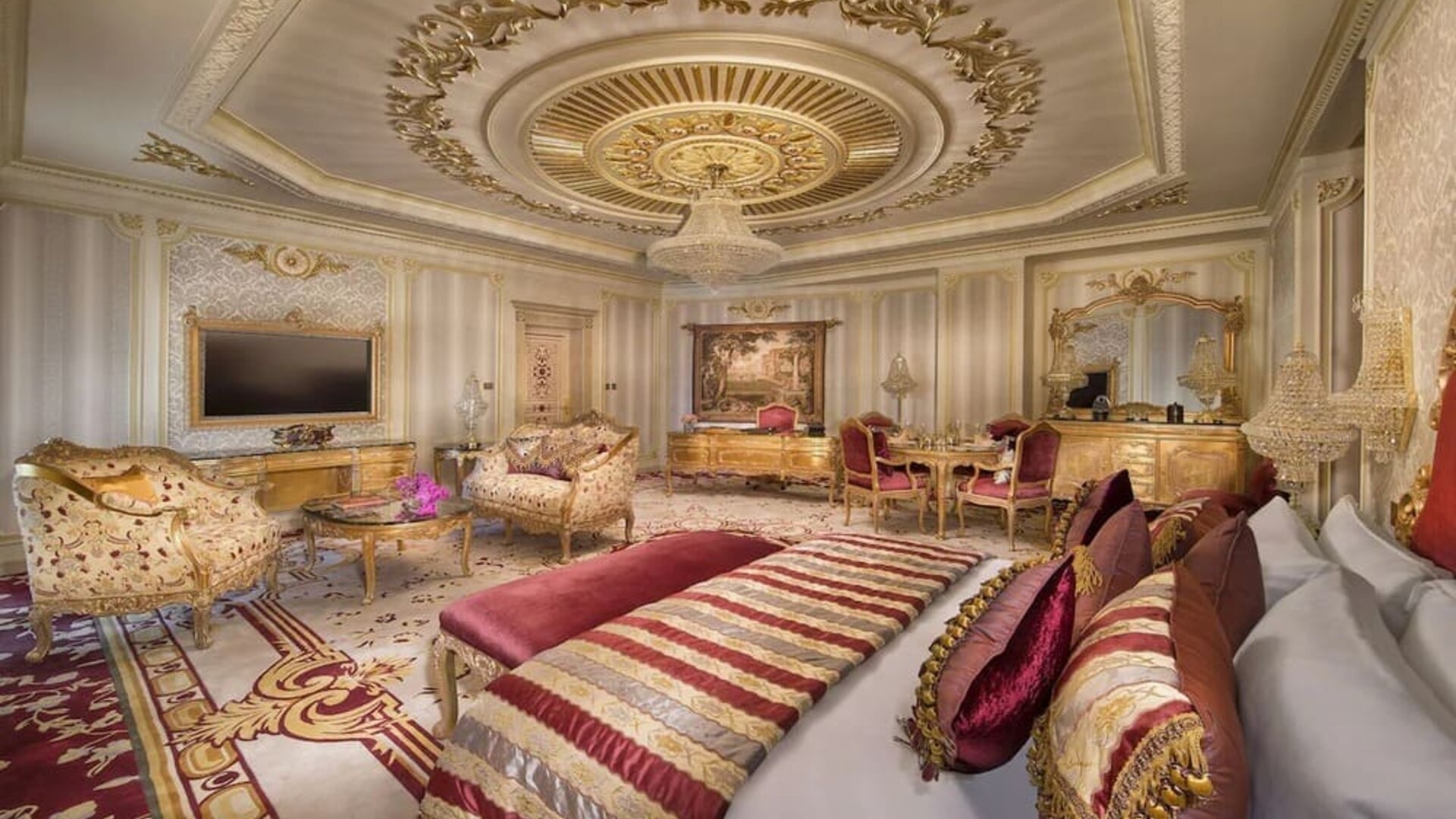 Luxurious suite with red and gold decor, ornate furniture, chandelier, and large bed