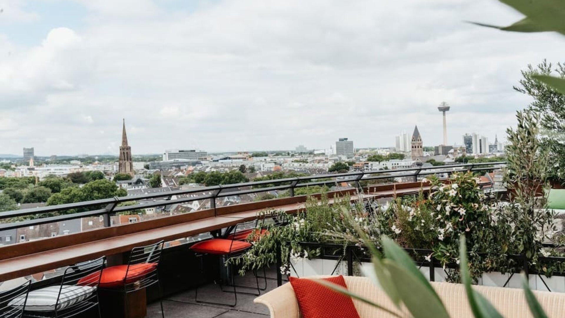 Rooftop terrace with cushioned seating, potted plants, and wide city skyline views beyond glass railing