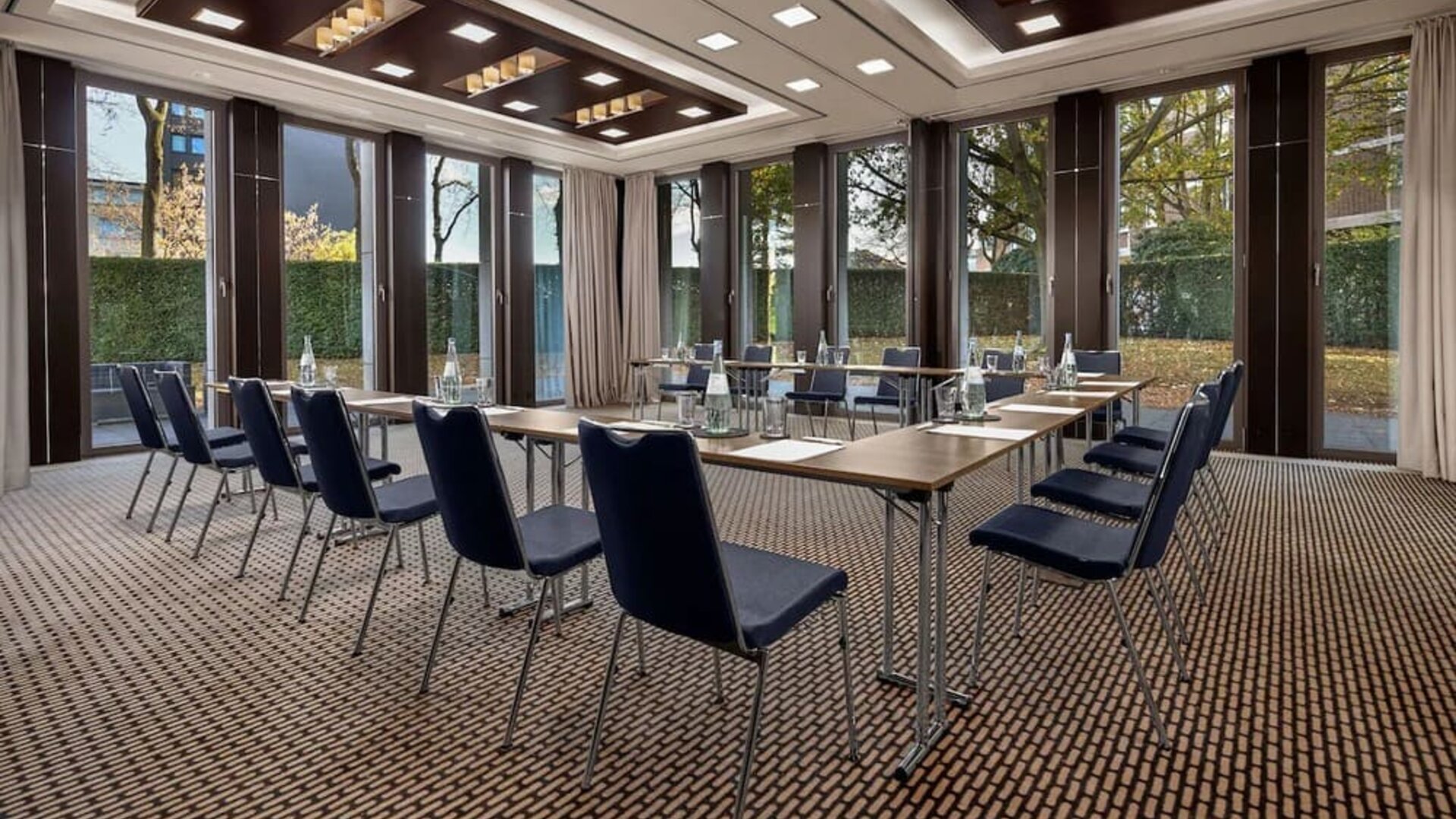 Meeting room with U-shaped tables, blue chairs, large windows, and garden view outside