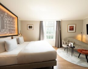 Cozy king bedroom with sofa seating area, wall-mounted TV, and large window framed by dark curtains
