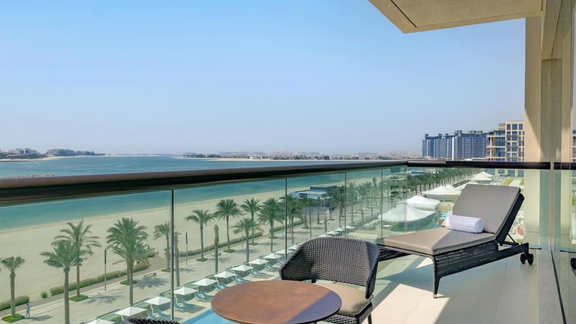 Balcony with lounge chair and table overlooking palm-lined beach, turquoise water, and distant city skyline