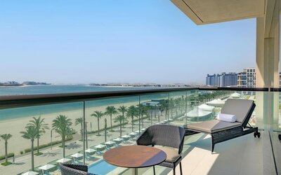 Balcony with lounge chair and table overlooking palm-lined beach, turquoise water, and distant city skyline