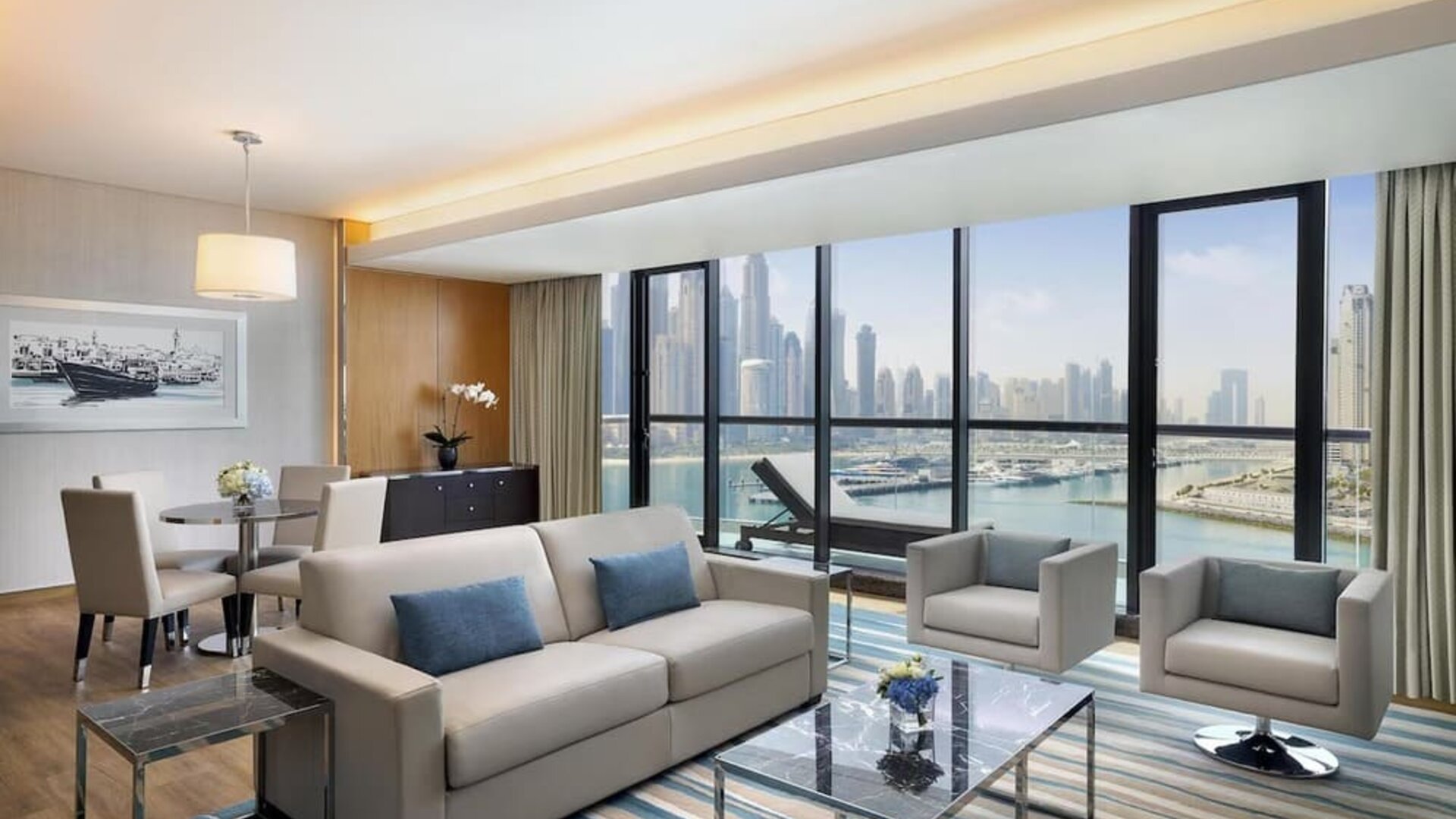 Living area with sofa, armchairs, dining table, and wide windows showcasing marina and skyline views