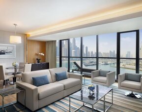 Living area with sofa, armchairs, dining table, and wide windows showcasing marina and skyline views