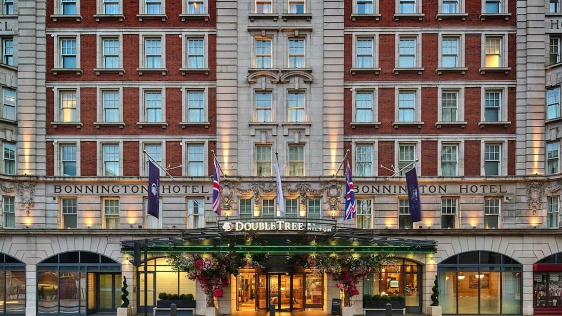 Red brick hotel exterior with lit entrance canopy, flags, and symmetrical rows of windows along the facade