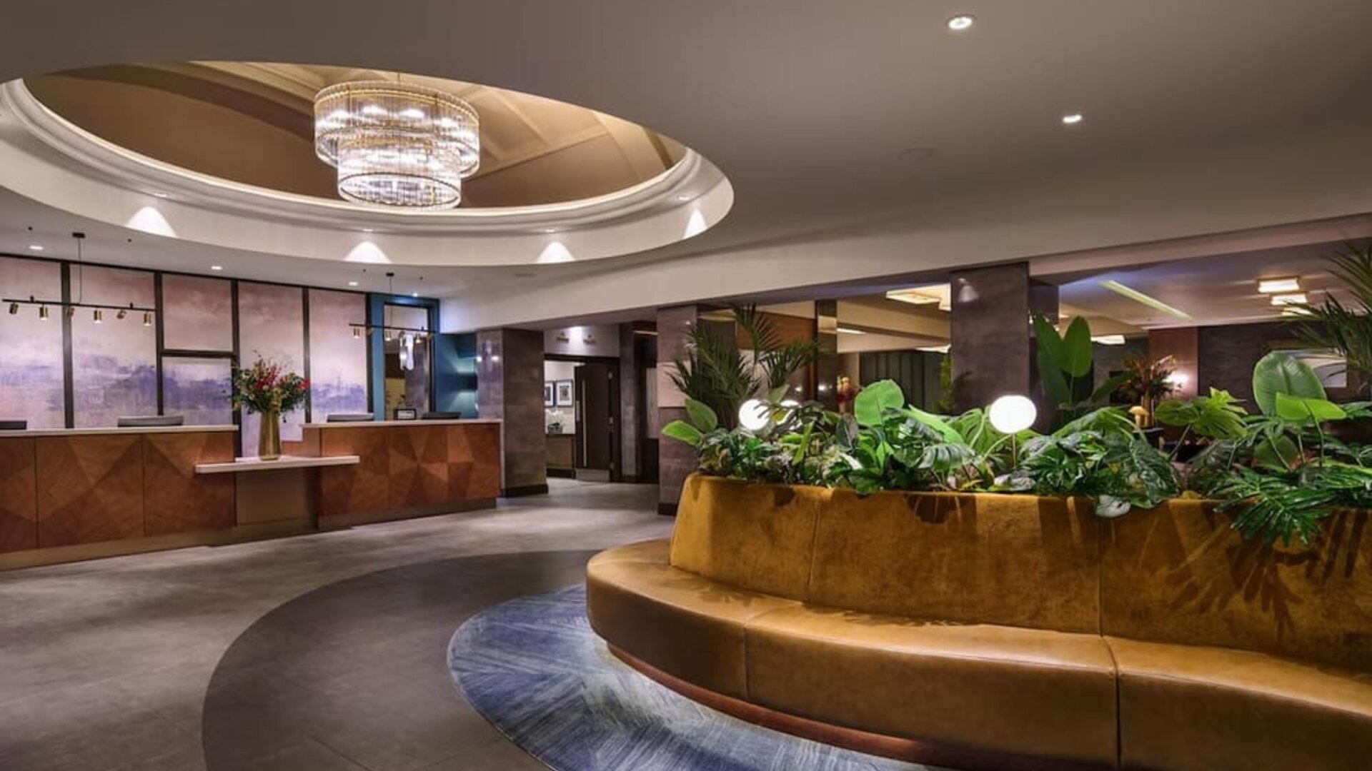 Hotel lobby with circular seating around indoor planter, chandelier above, and reception desk in background