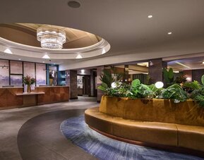 Hotel lobby with circular seating around indoor planter, chandelier above, and reception desk in background