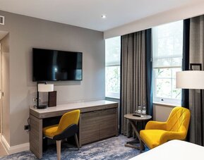 Desk with chair beneath wall-mounted TV, yellow armchair by window, and neutral tones with floor-length curtains