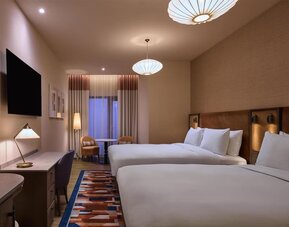Two queen beds with upholstered headboards, patterned rug, desk area, and seating nook near curtained window