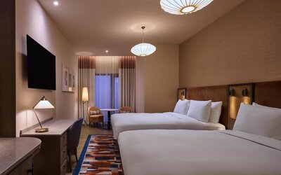 Two queen beds with upholstered headboards, patterned rug, desk area, and seating nook near curtained window