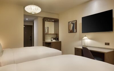 Two-bed guest room with desk, wall-mounted TV, and coffee station near entry in warm neutral tones