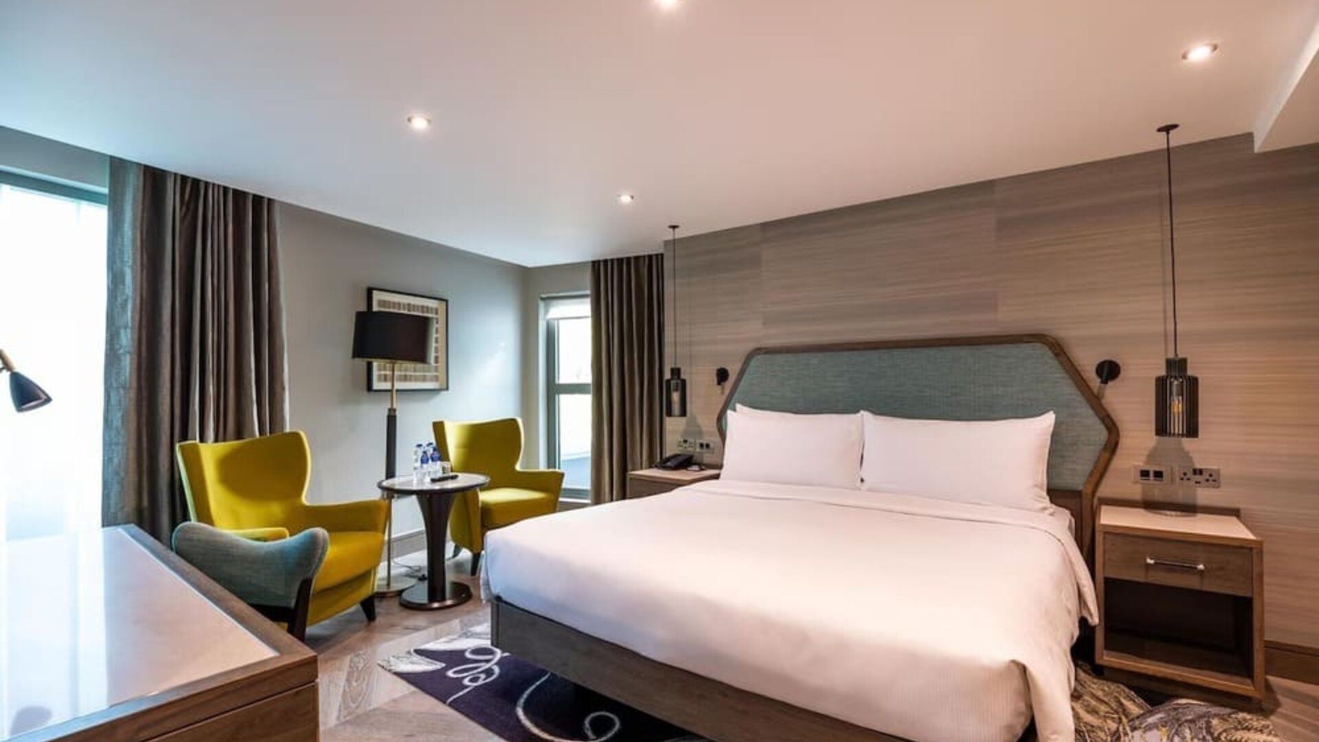 Spacious king room with large bed, padded headboard, desk, and seating area by tall window with dark curtains