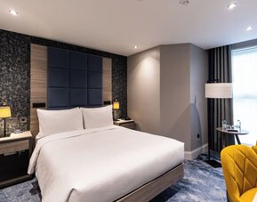 King bed with padded blue headboard, yellow chairs by window, and textured accent wall behind bedside tables