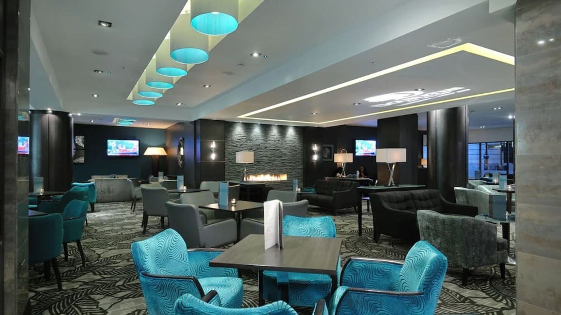 Modern lounge with teal chairs, patterned carpet, fireplace wall, and soft overhead lighting accents
