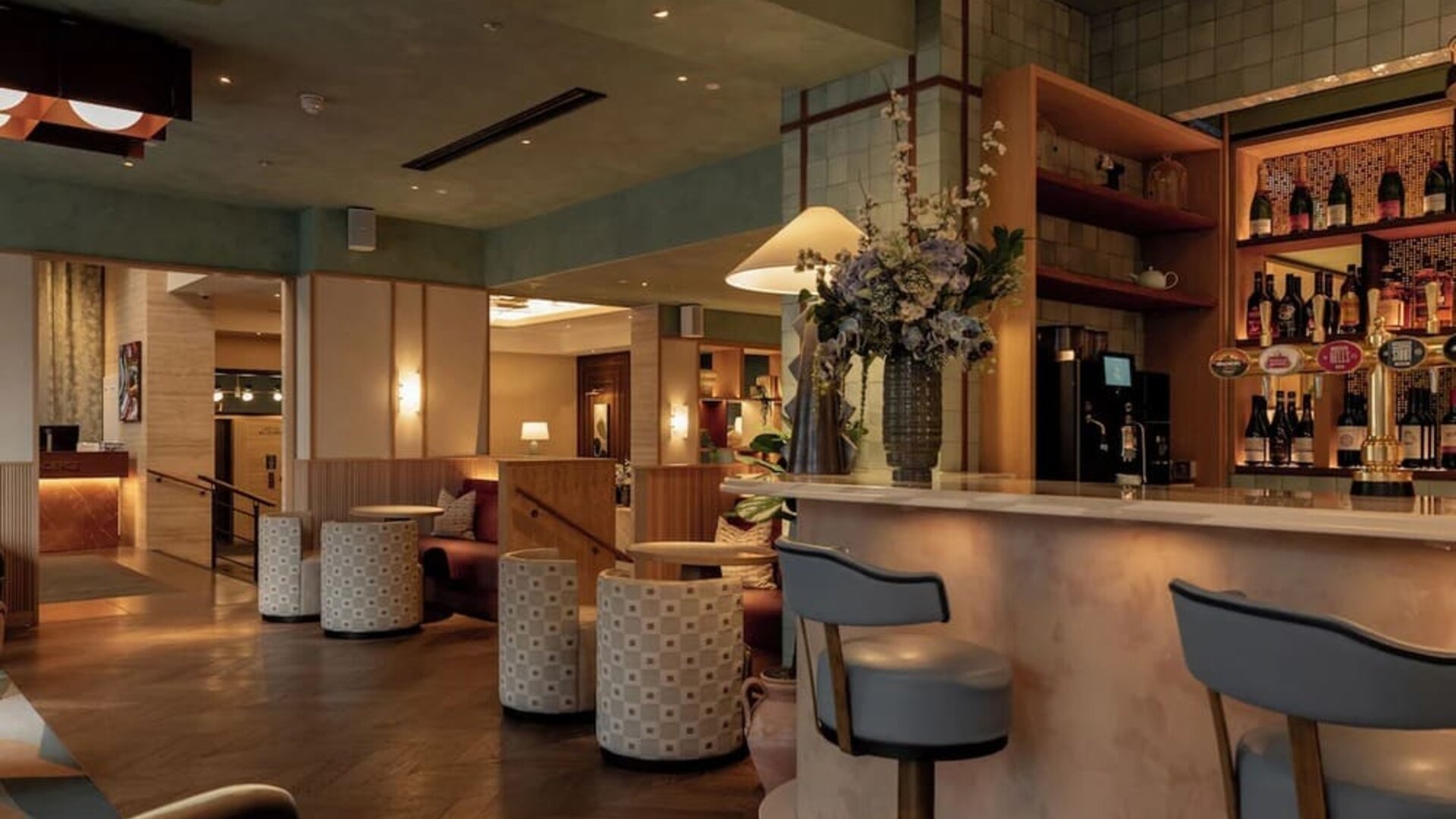 Hotel bar lounge with marble counter, upholstered stools, floral centerpiece, and warm lighting