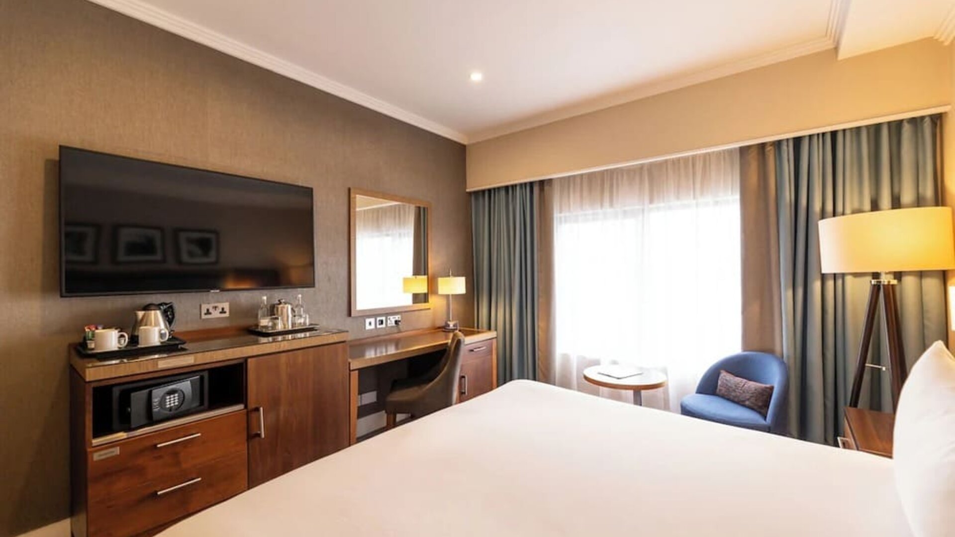 King guest room with wall-mounted TV, wood desk, blue accent chair, and large curtained window