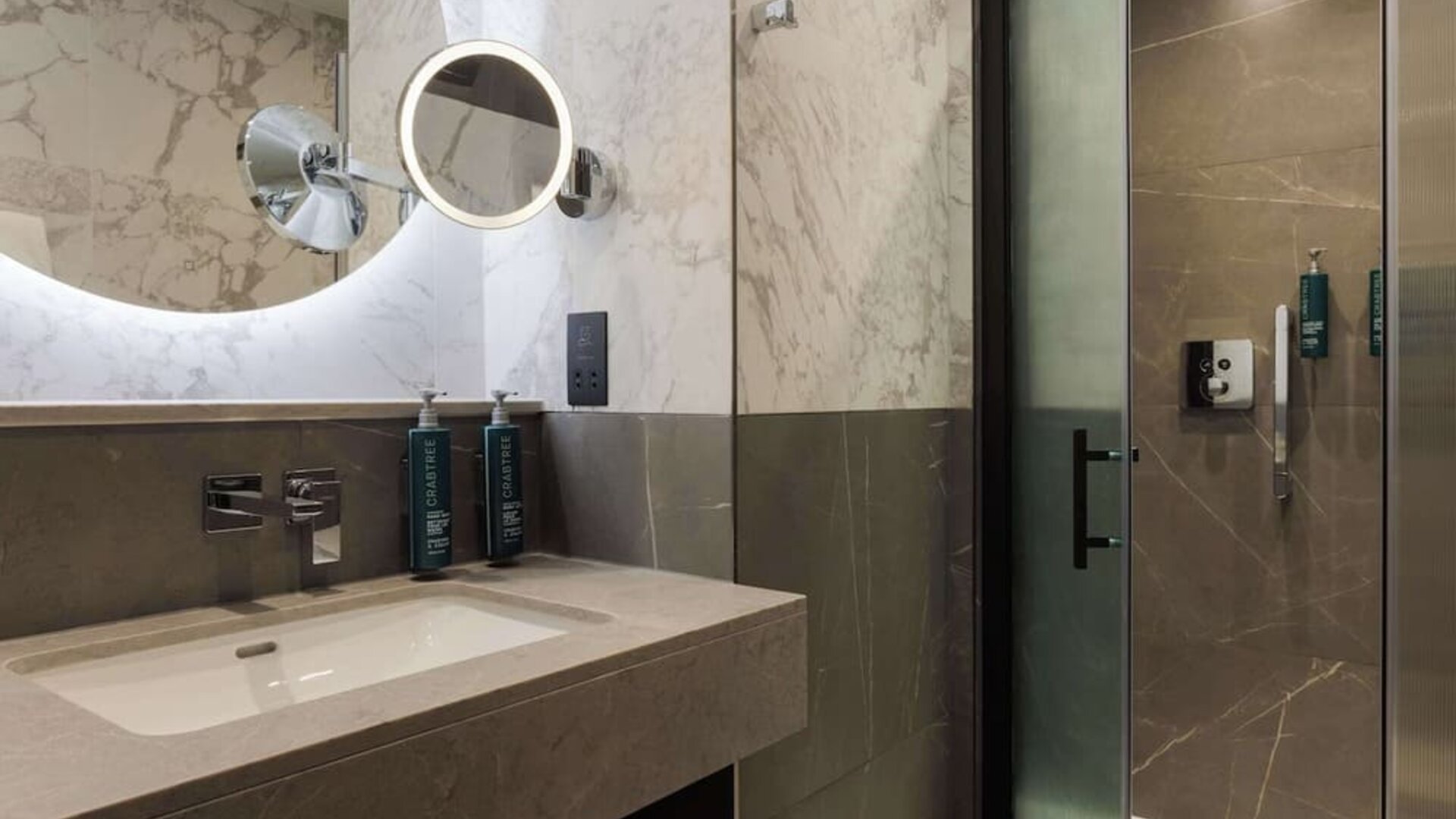 Marble bathroom with illuminated round mirror, modern sink vanity, and glass walk-in shower
