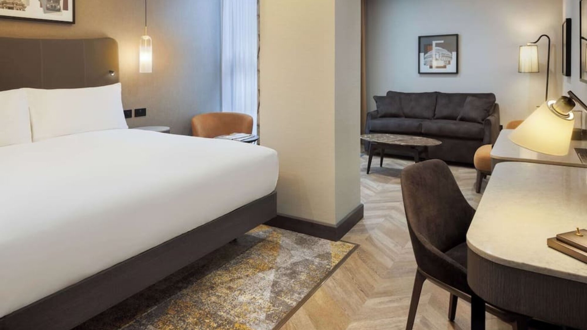 Suite-style king room with separate sitting area, sofa, curved desk, and modern lighting fixtures