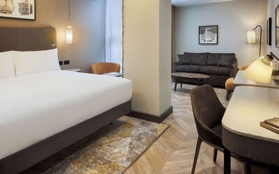 Suite-style king room with separate sitting area, sofa, curved desk, and modern lighting fixtures