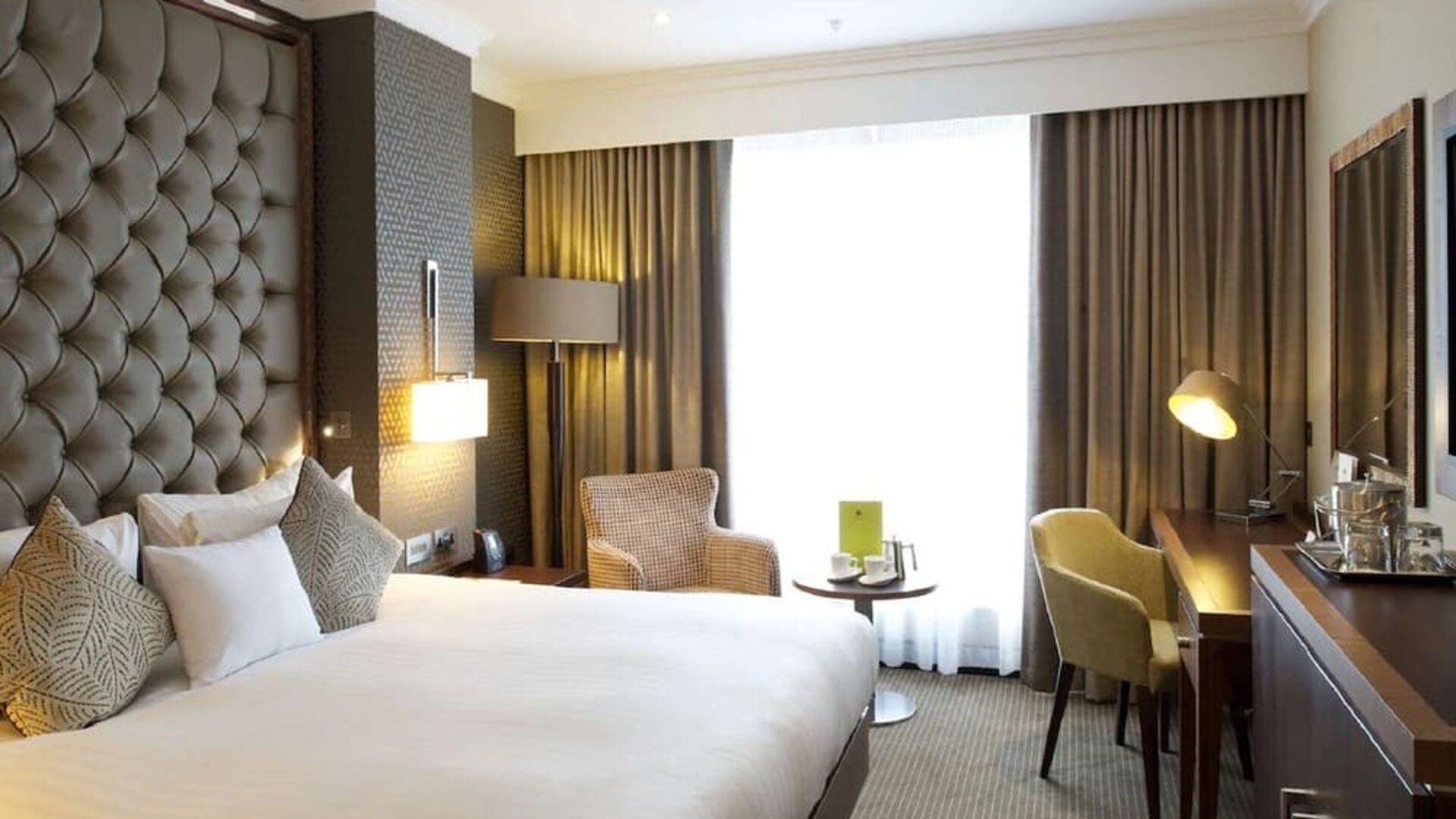 King bedroom with tall tufted headboard, armchair seating area, and desk beside bright window