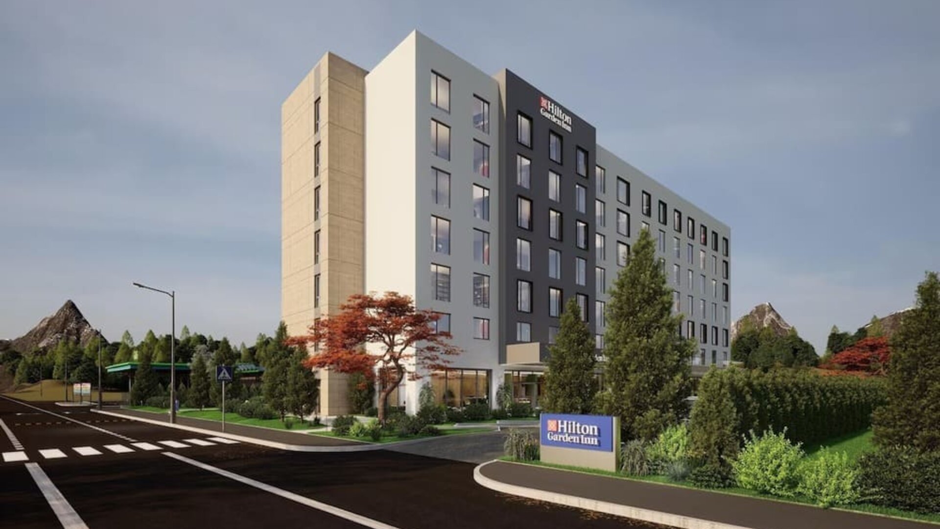 Modern seven-story hotel exterior with landscaped entrance, roadside sign, and trees at dusk