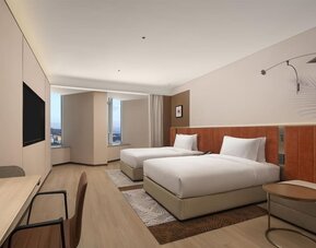 Modern guest room with two double beds, wall-mounted TV, desk area, and corner city windows