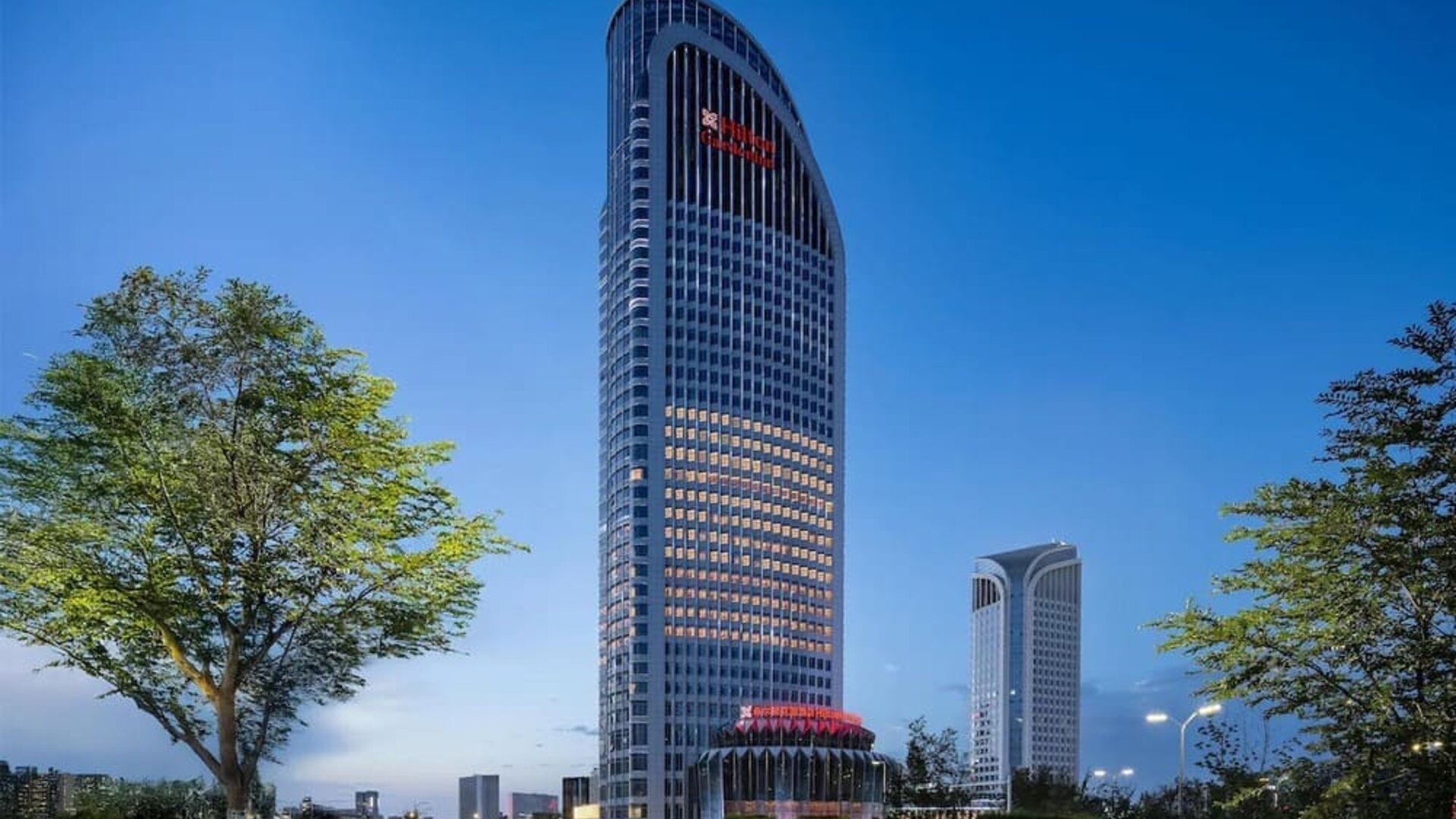 Tall curved hotel tower illuminated at dusk with landscaped grounds and city skyline in background