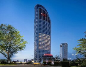 Tall curved hotel tower illuminated at dusk with landscaped grounds and city skyline in background