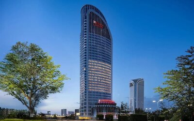 Tall curved hotel tower illuminated at dusk with landscaped grounds and city skyline in background