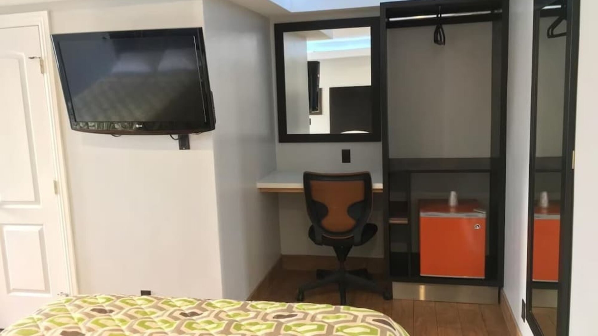 In-room workspace with desk, chair, mini fridge, wall-mounted TV, and open closet area at Mid City Inn - Los Angeles.
