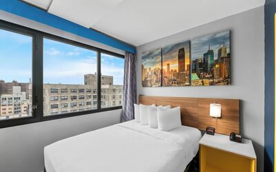 Queen bed beside large city-view window with skyline artwork and bright accents at Days Inn Chinatown NYC.