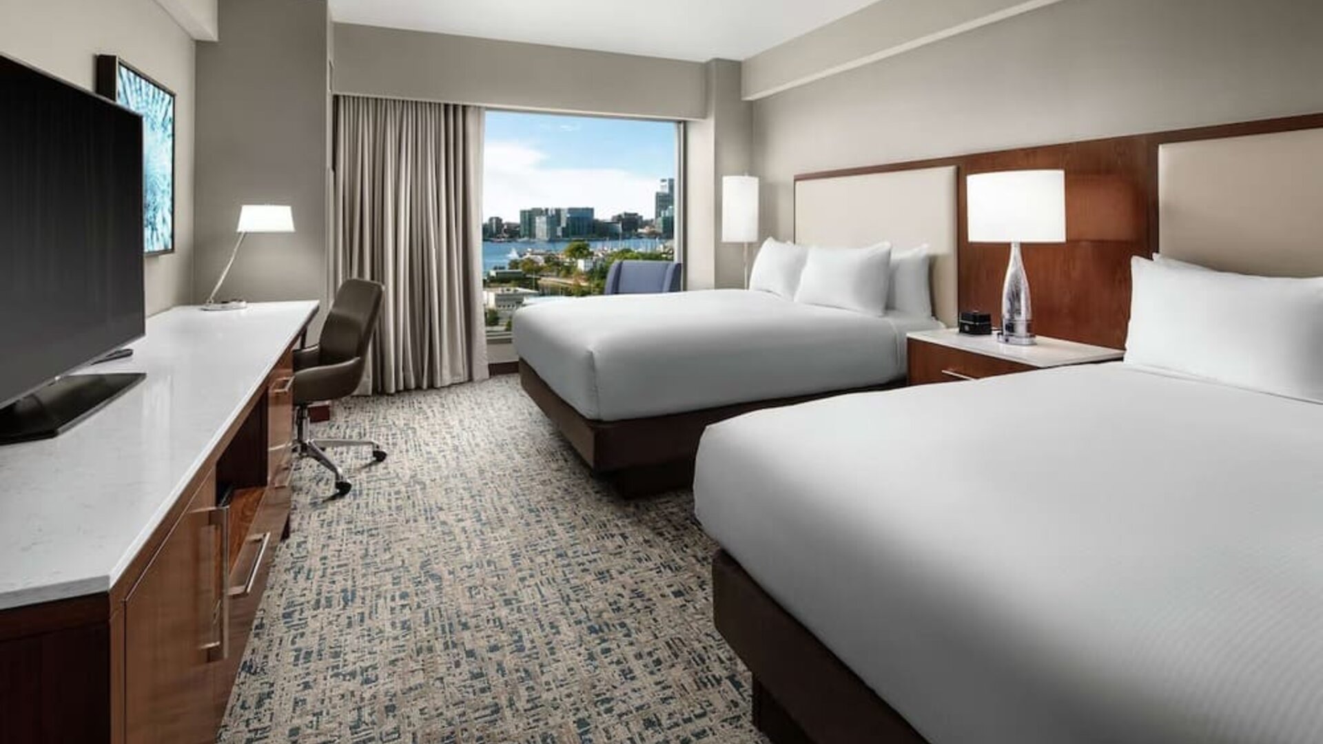 Room with two beds, workspace desk, and waterfront city view through large window at Hilton Boston Logan Airport.
