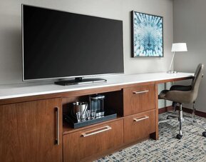 Workspace with desk chair, large TV, coffee station, and wooden cabinetry at Hilton Boston Logan Airport.