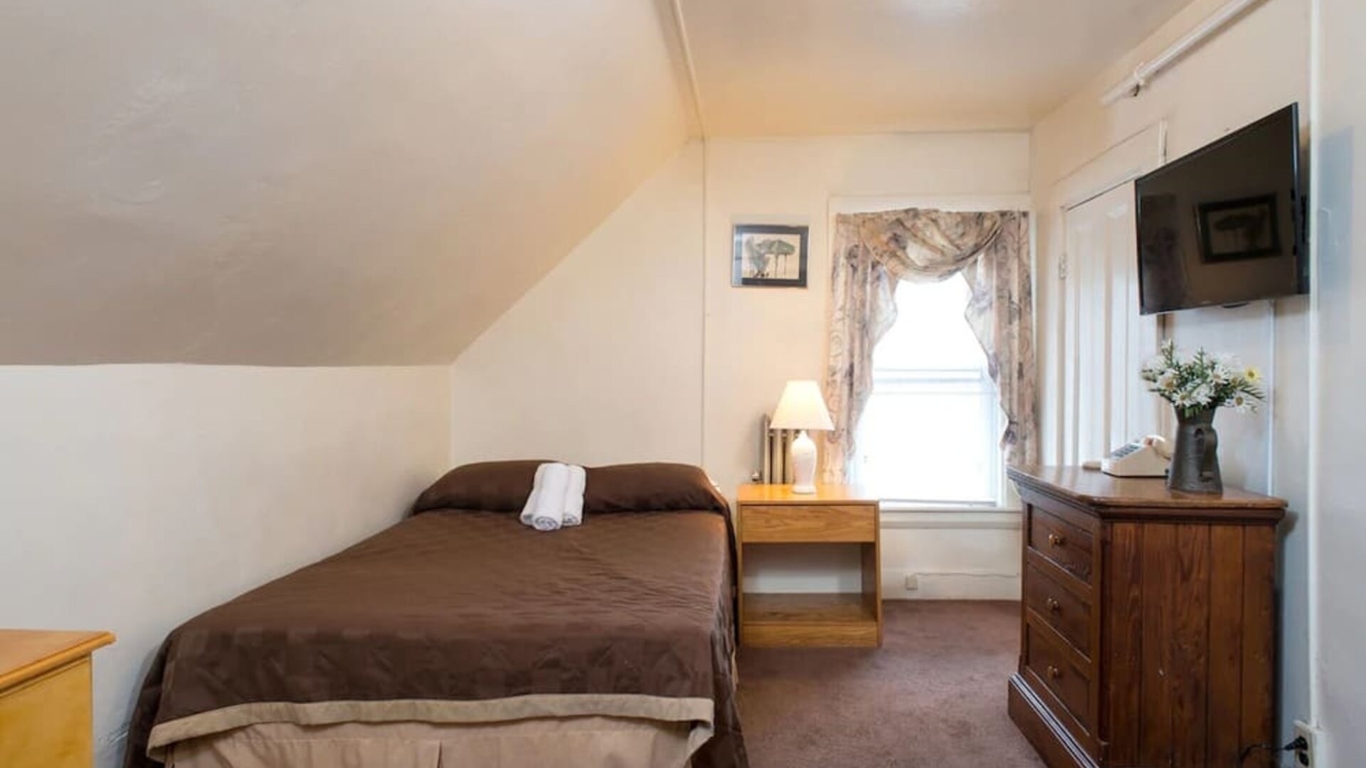 Small attic-style guest room with double bed, wooden dresser, and wall-mounted TV at The Farrington Inn.