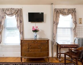 Seating and workspace area with wooden desk, dresser, floral curtains, and mounted TV at The Farrington Inn.