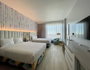 Spacious guest room with two beds, modern furnishings, oversized television, work area, and large windows providing abundant natural light.