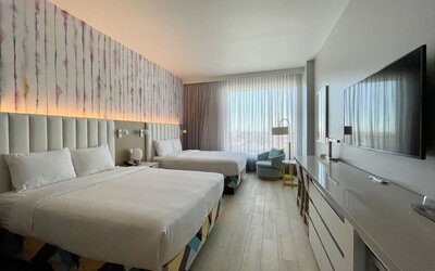 Spacious guest room with two beds, modern furnishings, oversized television, work area, and large windows providing abundant natural light.